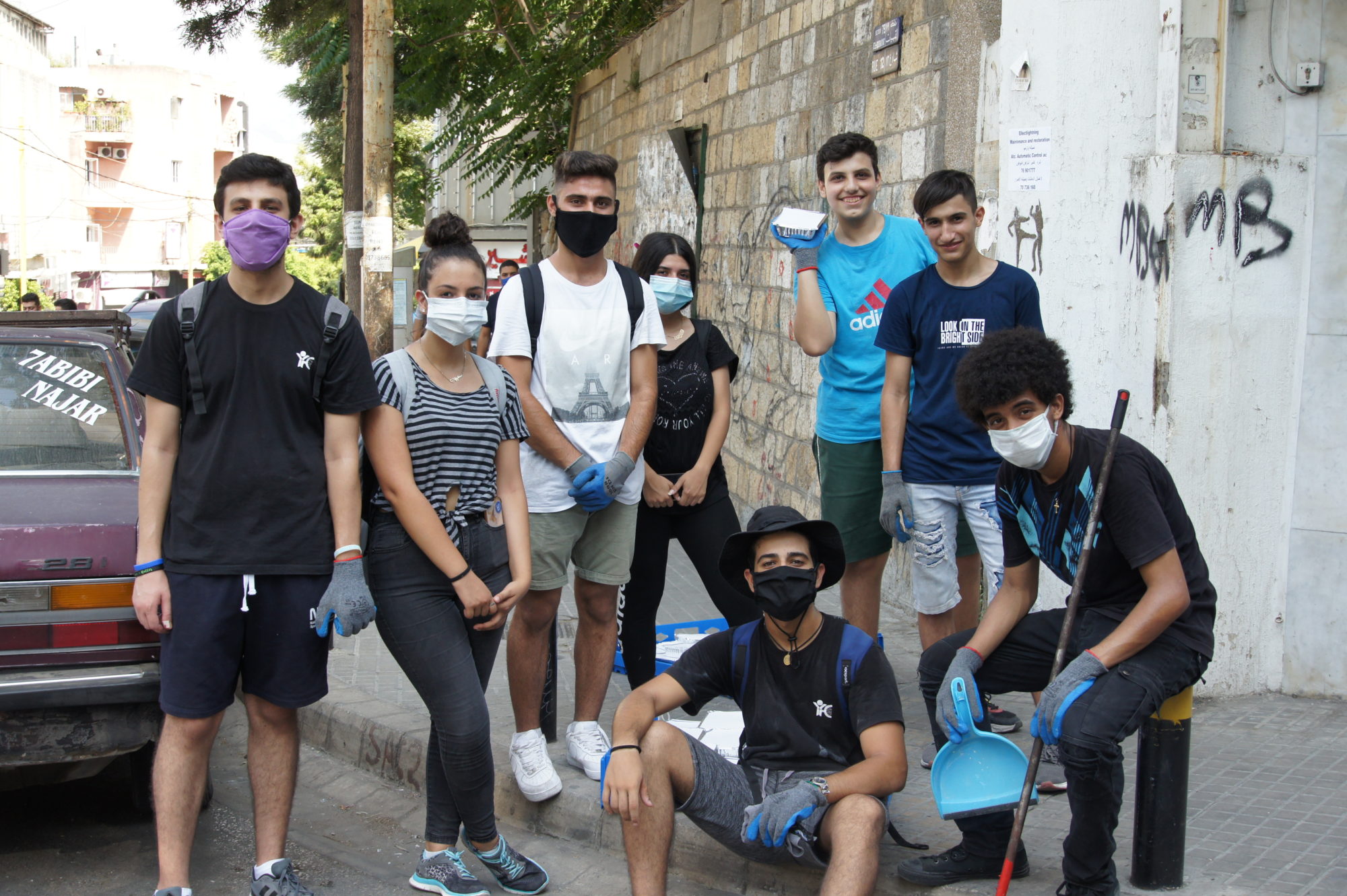Eight teenagers in Lebanon smile with cleaning supplies on a sidewalk