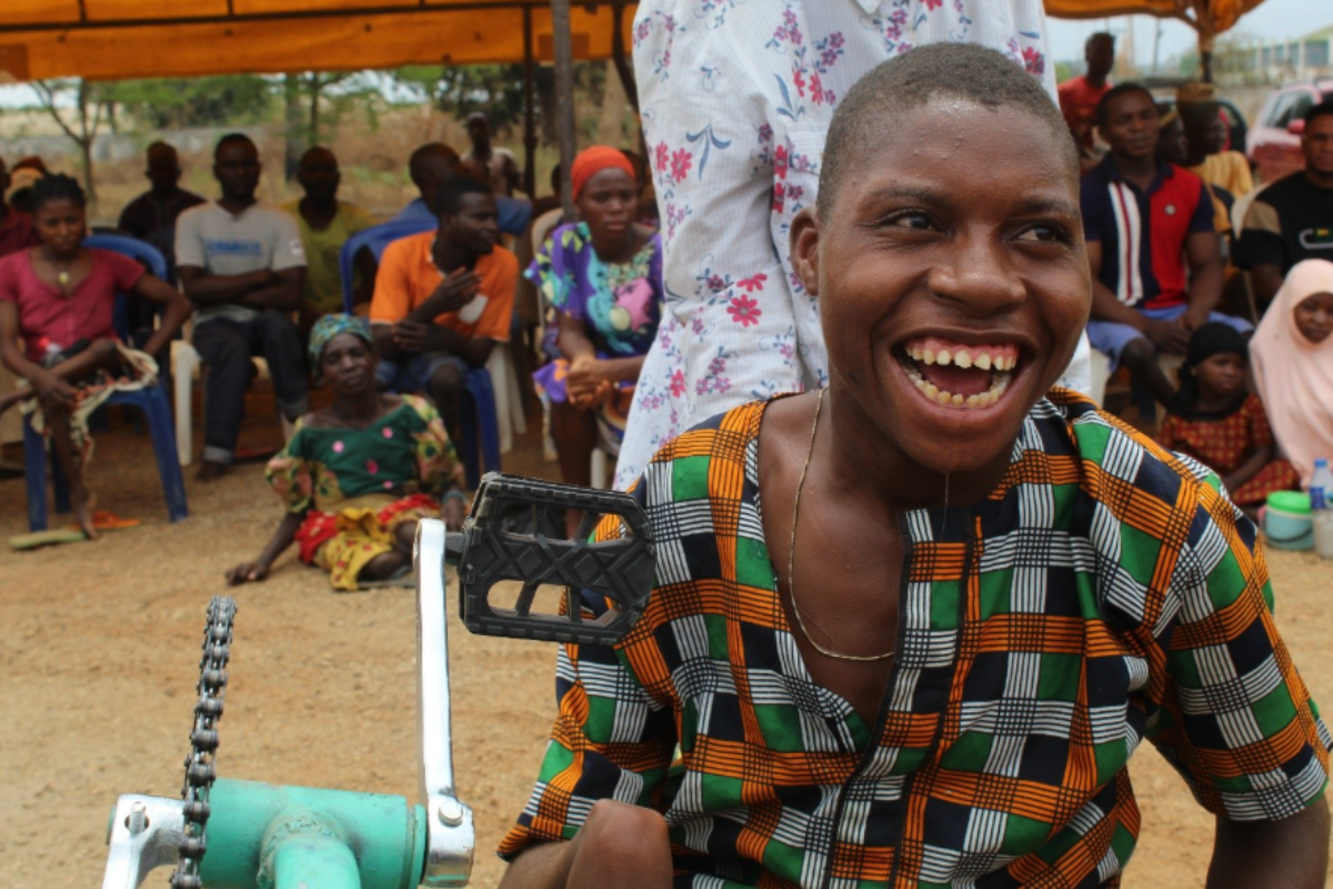 Person using a tricycle wheelchair outdoors with a group of seated people in the background.