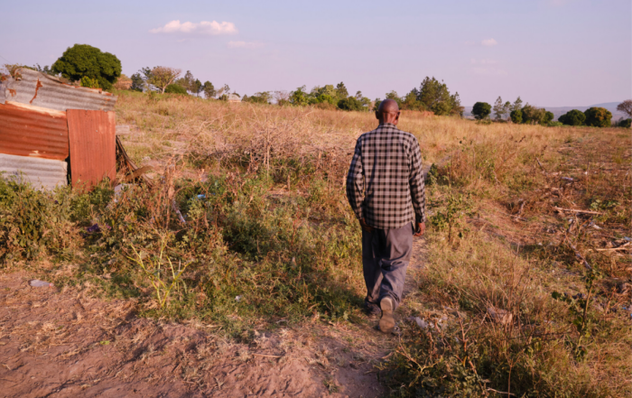 Person walking through a dry field near a small structure in a rural landscape with trees in the distance.