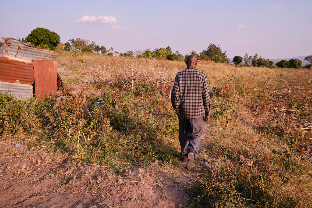 Person walking through a dry field near a small structure in a rural landscape with trees in the distance.
