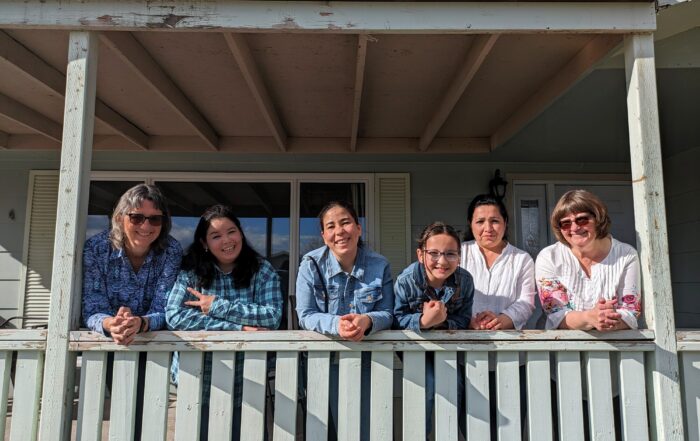 Six people leaning on a porch railing outside a house, gathered closely together in daylight.