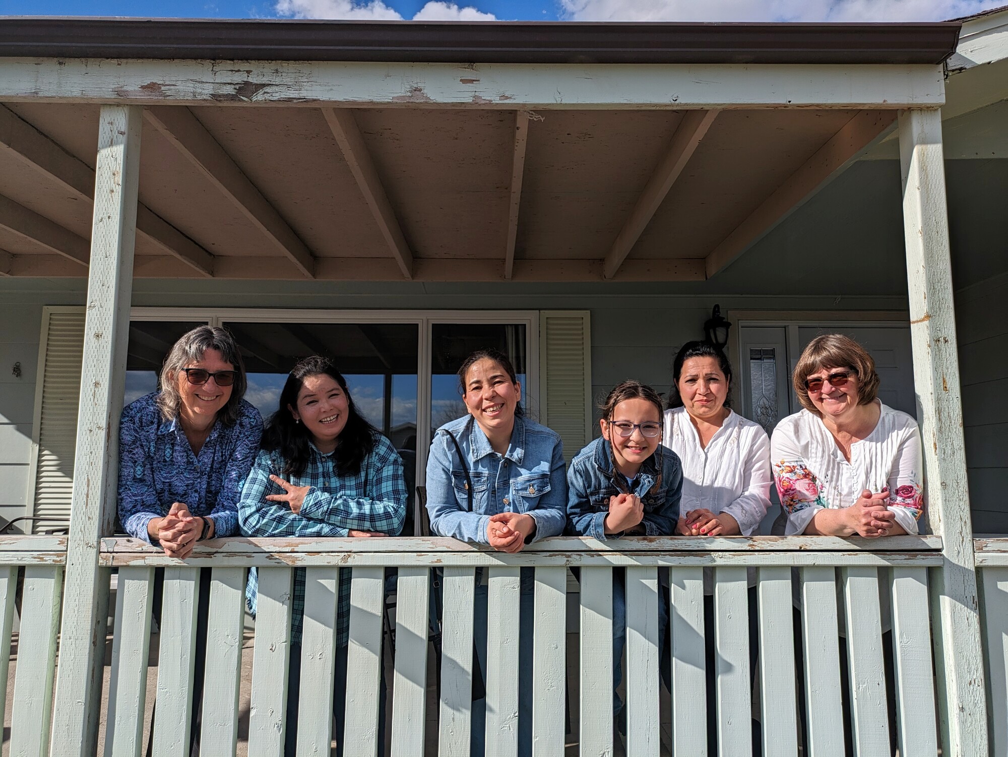Six people leaning on a porch railing outside a house, gathered closely together in daylight.